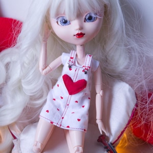 Op de afbeelding: Een pop met lang, wit haar en blauwe ogen, gekleed in een witte jurk met rode hartaccenten. De pop poseert op een wit kussen met een rood hartvormig kussen op de achtergrond. Een miniatuur rode viool ligt in de buurt.