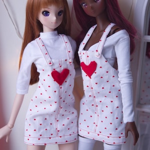 May include: Two dolls wearing white dresses with red heart patterns and red heart accents. The dresses have suspender straps and are paired with white long-sleeved shirts. One doll has auburn hair, and the other has dark red hair.