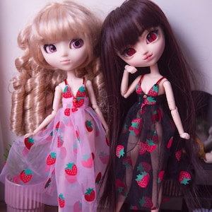 May include: Two fashion dolls wearing strawberry-print dresses. One doll has blonde curly hair and a pink dress, the other has dark hair and a black dress. Both dresses have red and green strawberry patterns.