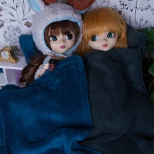 May include: Two dolls are lying in bed under blankets. One doll is wearing a white and brown hooded animal costume and holding a small cup. The other doll is wearing a long, orange wig and has green eyes.