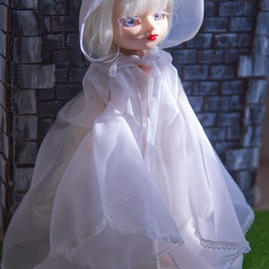 May include: A doll with pale skin, blue eyes, and red lips, wearing a white hooded cape and dress. The doll's blonde hair is visible under the hood. The cape and dress are made of a sheer fabric with lace trim. The doll is posed in front of a grey brick wall.