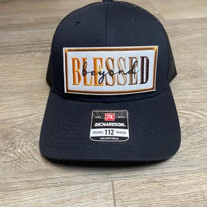 May include: Black trucker hat with a mesh back and a curved brim. The front features a rectangular patch with the word "BLESSED beyond" embroidered in gold and brown. The hat is adjustable and has a Richardson label.