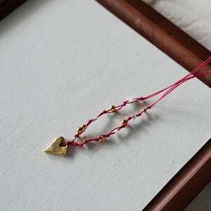 Könnte beinhalten: Eine Halskette mit einem goldenen Herz-Anhänger, roter Schnur und goldenen Perlen. Die Halskette wird auf einem Tablett aus Holz mit einer weißen Oberfläche präsentiert.