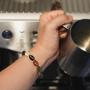 Könnte beinhalten: Eine Hand hält einen Milchaufschäumer aus Edelstahl. Der Milchaufschäumer wird über einer Kaffeemaschine gehalten. Die Person trägt ein Armband mit bunten Perlen.