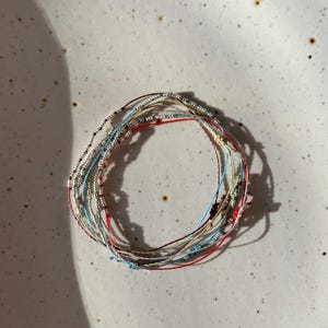 Puede incluir: Una colección de delicadas pulseras con cuentas en varios colores, incluyendo plata, rojo, azul y beige. Las pulseras están hechas con cordones finos y pequeñas cuentas, creando un aspecto minimalista y elegante. Las pulseras están dispuestas en forma circular.