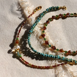 May include: Three beaded bracelets with gold clasps. The top bracelet is turquoise blue, the middle bracelet is brown and green, and the bottom bracelet is red and gold.