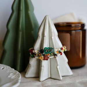 May include: Three colorful beaded bracelets with green, red, and gold accents are displayed on a white ceramic Christmas tree ornament. The ornament is sitting on a beige fabric surface.