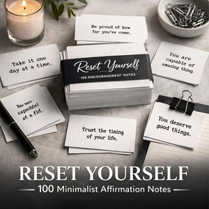 May include: A stack of white affirmation cards with the text "Reset Yourself" and "100 Minimalist Affirmation Notes". Individual cards with motivational quotes are scattered around, along with a lit candle and a pen.