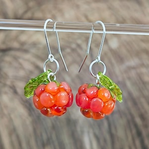May include: A pair of handcrafted earrings featuring clusters of orange and red beads, resembling raspberries. Each earring has a small green leaf accent and hangs from a silver-colored hook.