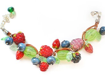 Op de afbeelding: Een armband gemaakt van bruin lederen koord met glazen kralen in de vorm van frambozen, bosbessen en aardbeien. De kralen zijn groen, rood, roze en blauw.
