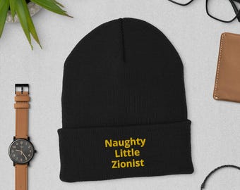 Może przedstawiać: Czarna czapka z napisem "Naughty Little Zionist" wyhaftowanym na żółto. Czapka jest złożona na brzegu. Widoczny jest również zegarek, okulary i portfel.