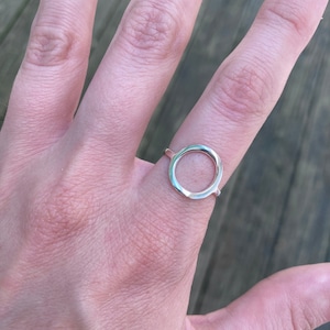 May include: A silver ring with a simple, open circle design. The ring is worn on a person's finger.