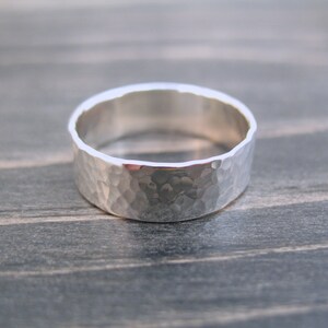May include: A wide, hammered silver ring. The ring has a textured surface with a series of small indentations, giving it a unique, handcrafted appearance. The band is approximately 0.3 inches wide. The ring is set against a neutral background.