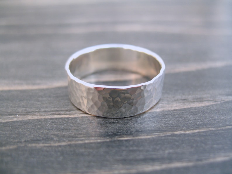 May include: A wide, hammered silver ring. The ring has a textured surface with a series of small indentations, giving it a unique, handcrafted appearance. The band is approximately 0.3 inches wide. The ring is set against a neutral background.