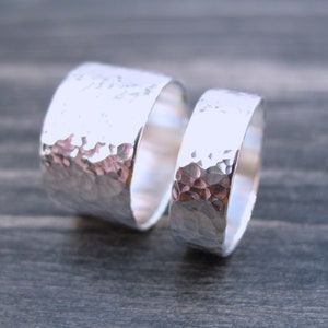 May include: Two silver rings with a hammered texture. The rings are different sizes, with one wider than the other. The rings are displayed on a dark gray wooden surface. The rings are likely for a couple.