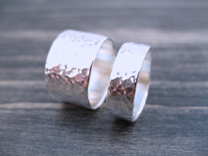 May include: Two silver rings with a hammered texture. The rings are different sizes, with one wider than the other. The rings are displayed on a dark gray wooden surface. The rings are likely for a couple.