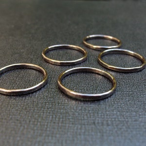 May include: Five simple gold rings, each with a hammered finish, are arranged on a dark gray surface.