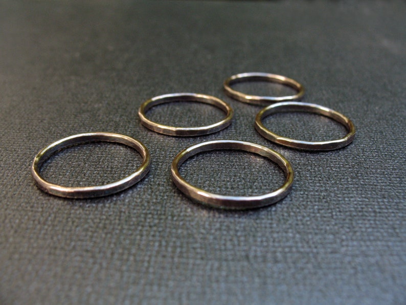 May include: Five simple gold rings, each with a hammered finish, are arranged on a dark gray surface.