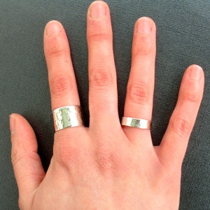 May include: Two silver rings are displayed on a hand. One ring is wide with a hammered texture, and the other is a thinner, smooth band. The rings are made of a shiny, metallic material.