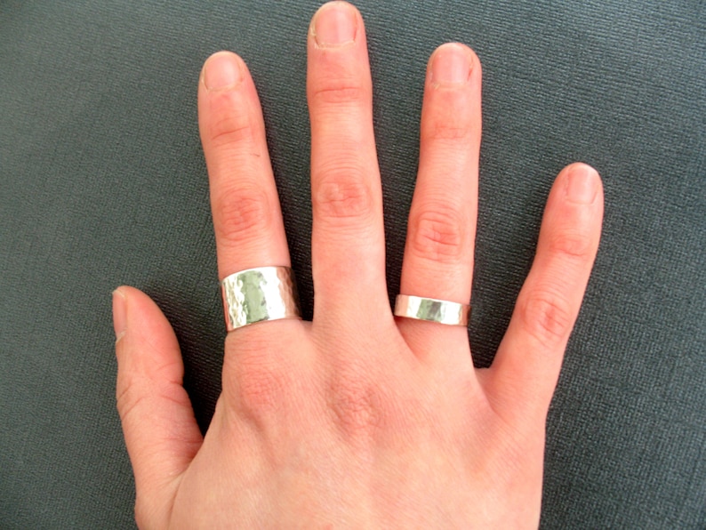 May include: Two silver rings are displayed on a hand. One ring is wide with a hammered texture, and the other is a thinner, smooth band. The rings are made of a shiny, metallic material.