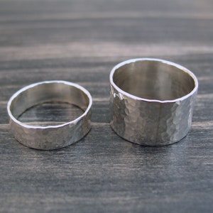 May include: Two silver rings with a hammered texture. One ring is wider than the other. The rings are displayed on a dark wooden surface. The rings are likely for a wedding or engagement.