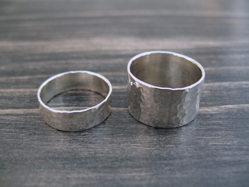 May include: Two silver rings with a hammered texture. One ring is wider than the other. The rings are displayed on a dark wooden surface. The rings are likely for a wedding or engagement.