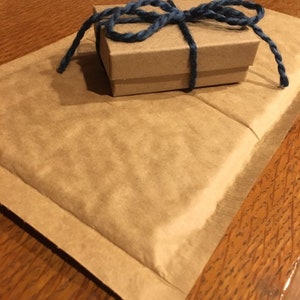 May include: A small, brown cardboard gift box tied with a dark blue twine bow, resting on a folded brown paper envelope. The items are on a wooden surface, creating a rustic aesthetic.