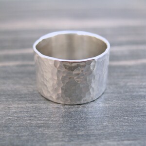 May include: A wide, silver ring with a hammered texture. The ring has a simple, minimalist design and a polished interior. The band is approximately 0.5 inches wide.