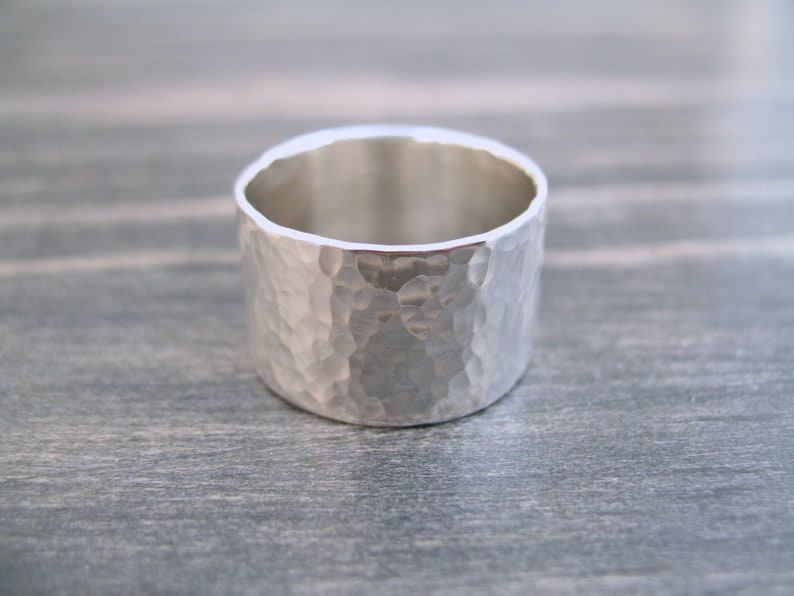 May include: A wide, silver ring with a hammered texture. The ring has a simple, minimalist design and a polished interior. The band is approximately 0.5 inches wide.