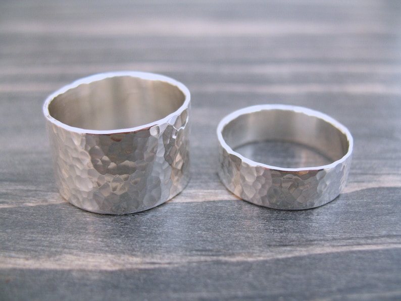 May include: Two hammered silver rings. The larger ring has a wider band and a textured surface. The smaller ring has a narrower band with a similar hammered texture. Both rings are displayed on a gray wooden surface.