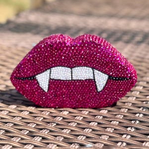 May include: A decorative, fuchsia-colored lips-shaped object, encrusted with sparkling crystals. The lips feature white beaded teeth and black fangs, creating a vampiric aesthetic. The object is placed on a woven surface.
