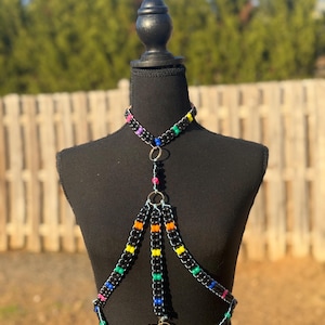 May include: A black beaded body chain with colorful accents. The chain features multiple strands with small, square-shaped beads. The chain has a choker style neckline and a decorative ring at the bottom.