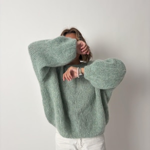 May include: A light green, oversized sweater with voluminous sleeves. The sweater has a textured knit design and is paired with white pants. The overall style is casual and comfortable, suitable for cooler weather.