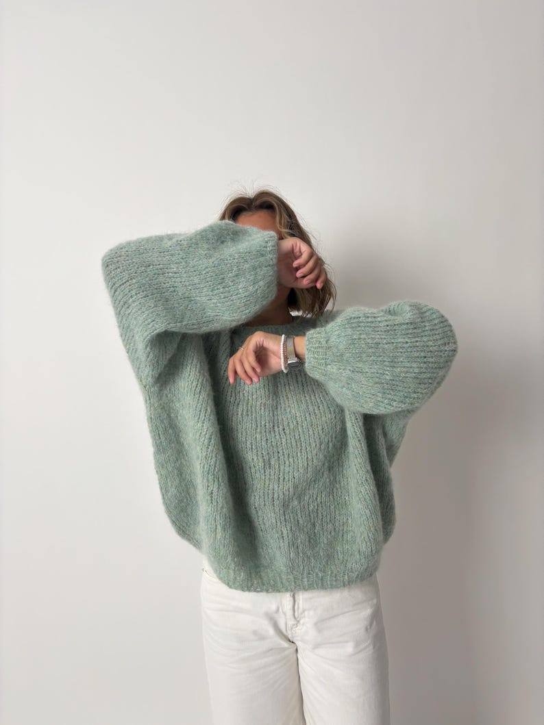 May include: A light green, oversized sweater with voluminous sleeves. The sweater has a textured knit design and is paired with white pants. The overall style is casual and comfortable, suitable for cooler weather.