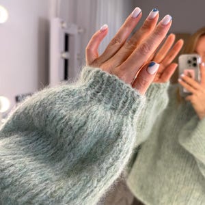 May include: A light green, fluffy knit sweater with a ribbed cuff. The image shows a close-up of the sleeve and cuff. The nails are painted white with a blue accent. The sweater appears soft and comfortable.