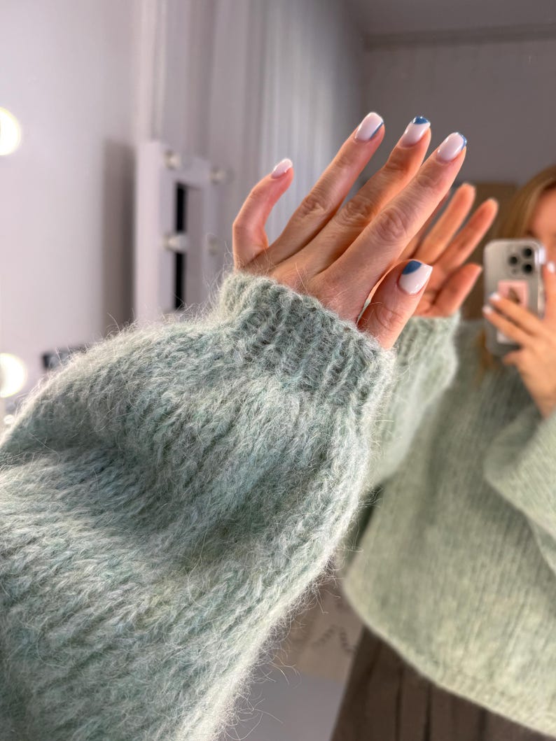 May include: A light green, fluffy knit sweater with a ribbed cuff. The image shows a close-up of the sleeve and cuff. The nails are painted white with a blue accent. The sweater appears soft and comfortable.