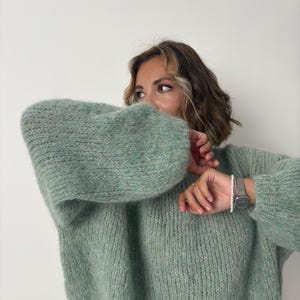 May include: A light green, oversized knit sweater with long, voluminous sleeves. The sweater has a soft, textured appearance and a relaxed fit. A silver watch with a white beaded bracelet is visible on the wrist.