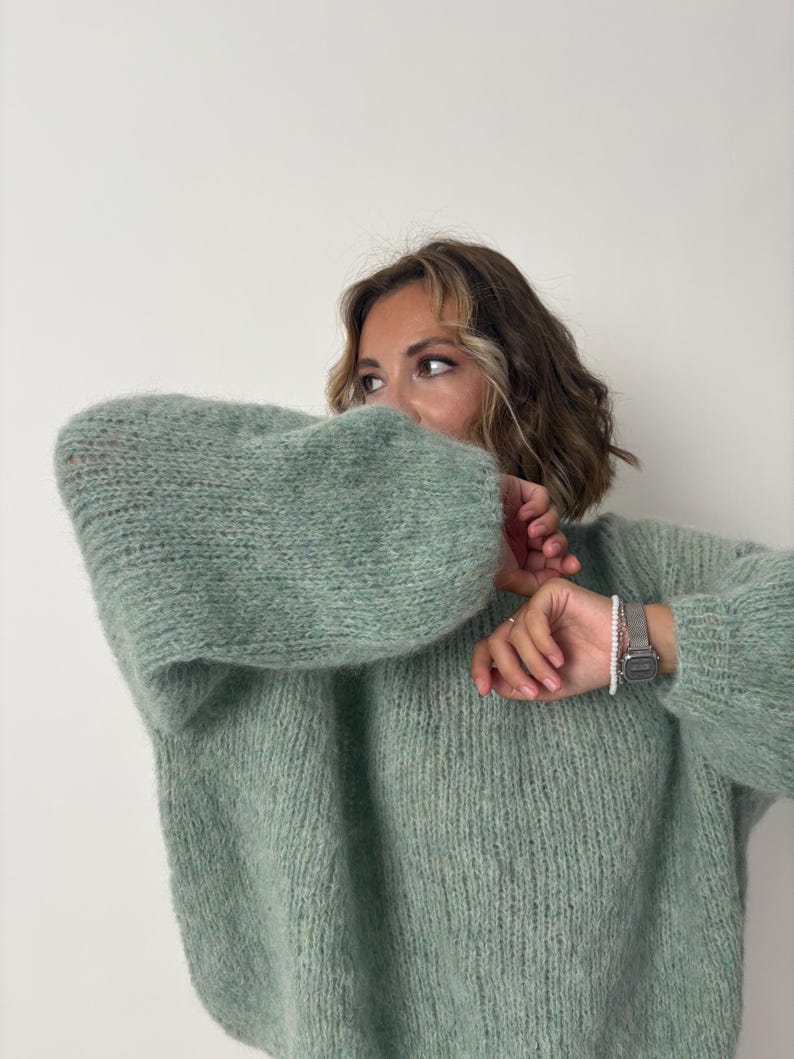 May include: A light green, oversized knit sweater with long, voluminous sleeves. The sweater has a soft, textured appearance and a relaxed fit. A silver watch with a white beaded bracelet is visible on the wrist.