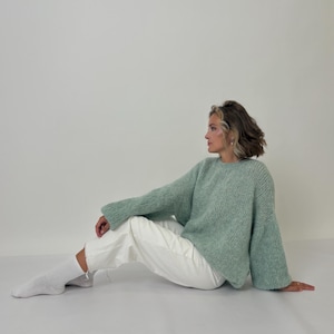 May include: A light green, oversized sweater with long sleeves and a crew neck. The sweater is paired with white pants and white socks. The model is sitting on a white surface.