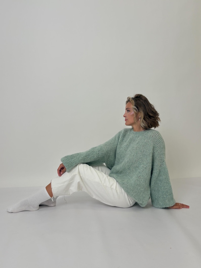May include: A light green, oversized sweater with long sleeves and a crew neck. The sweater is paired with white pants and white socks. The model is sitting on a white surface.