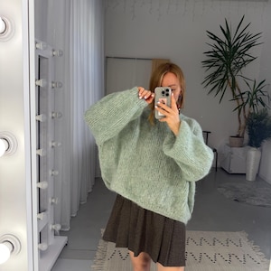 May include: A light green, oversized knit sweater with long sleeves. The sweater is paired with a brown pleated skirt and white socks. The image is taken in front of a mirror with a light fixture.
