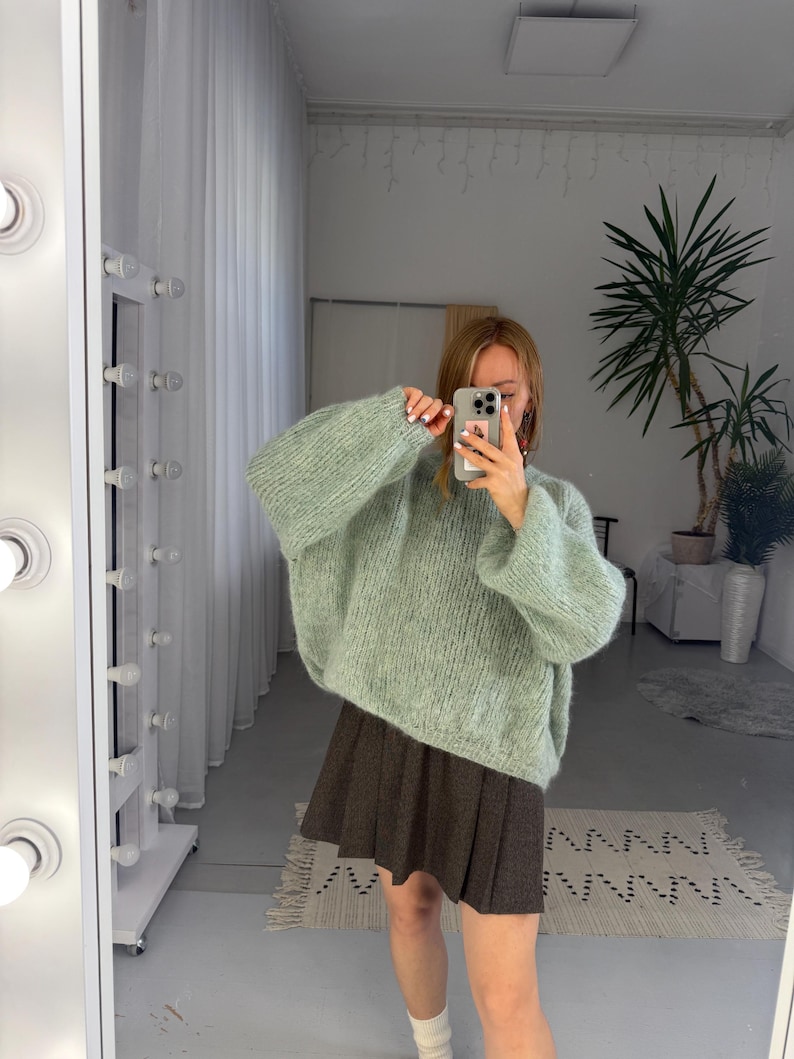 May include: A light green, oversized knit sweater with long sleeves. The sweater is paired with a brown pleated skirt and white socks. The image is taken in front of a mirror with a light fixture.