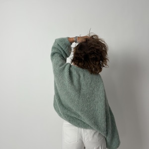 May include: A light green, oversized knit sweater with long sleeves. The sweater is paired with white pants. The person is facing away from the camera, with their hand in their hair. The background is a plain white wall.