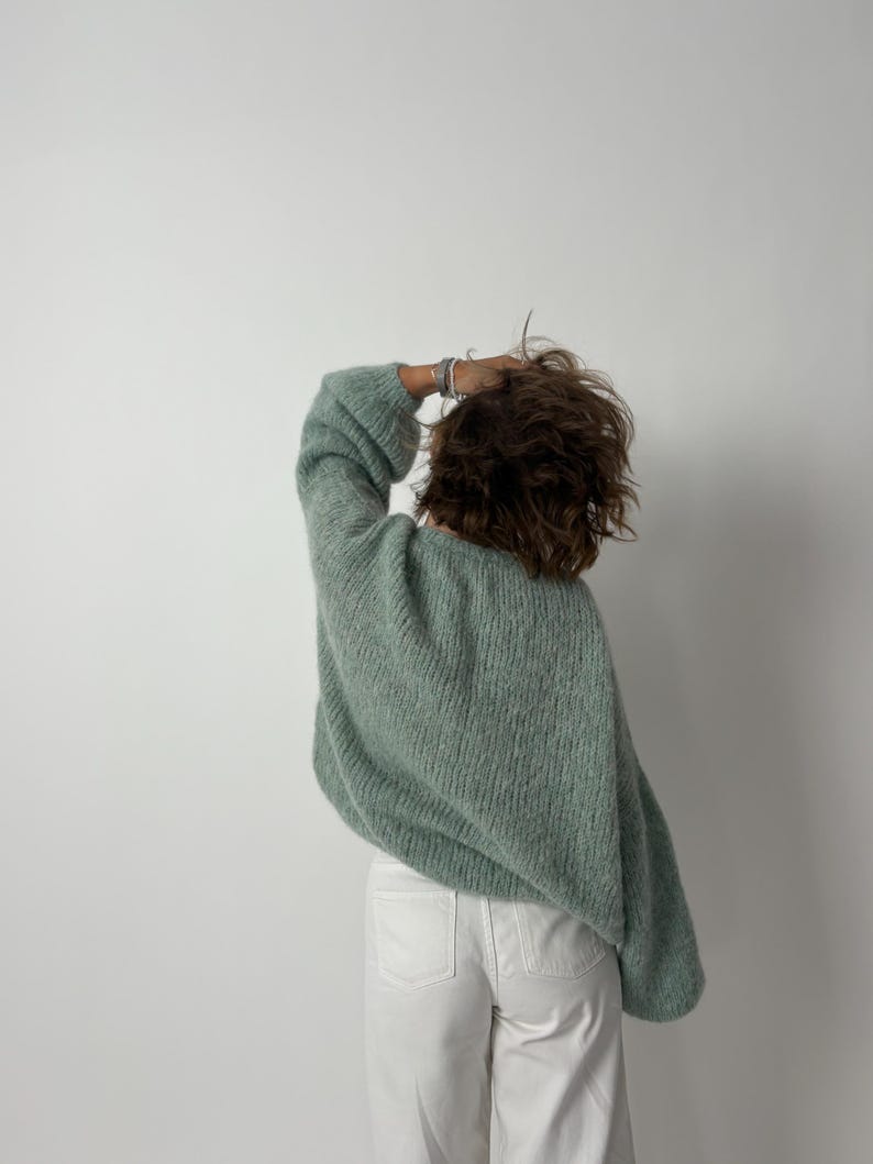 May include: A light green, oversized knit sweater with long sleeves. The sweater is paired with white pants. The person is facing away from the camera, with their hand in their hair. The background is a plain white wall.