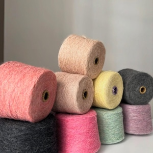 May include: A collection of colorful yarn spools in various shades, including pink, peach, yellow, green, and gray. The spools are stacked, showcasing the texture and color variations of the yarn. Ideal for knitting and crochet projects.