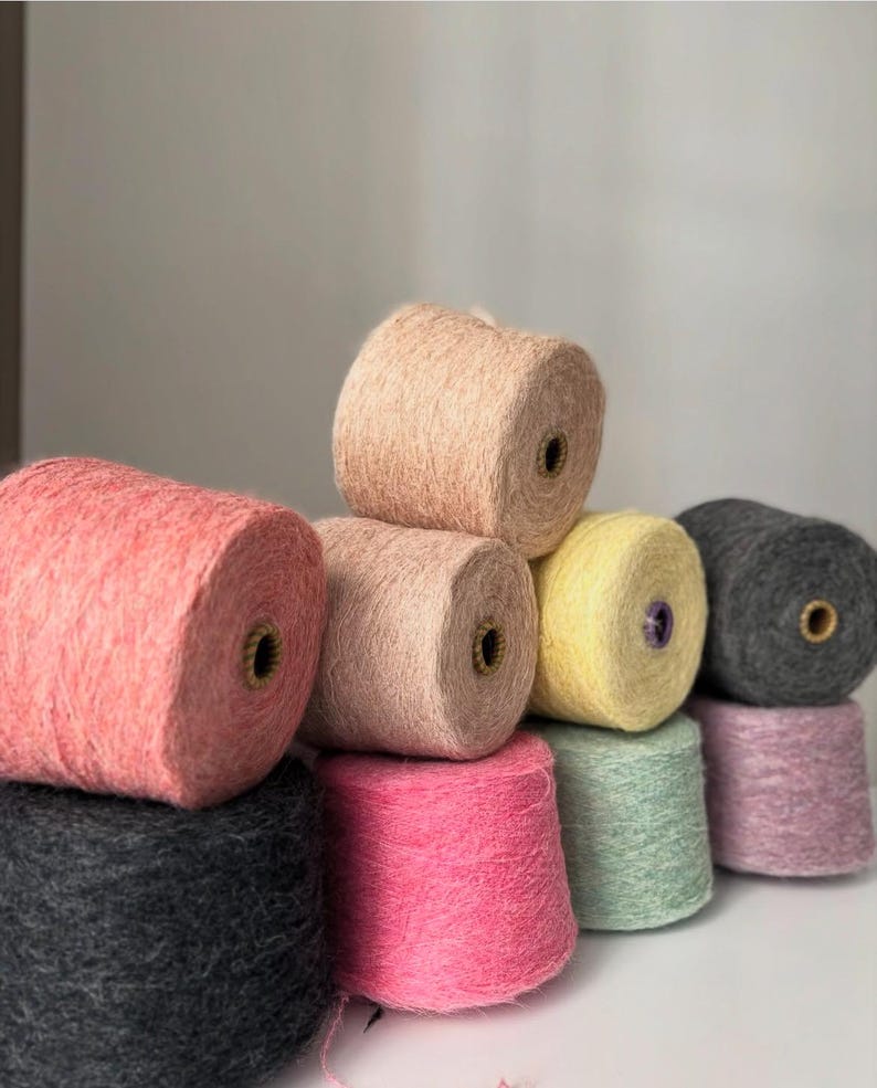 May include: A collection of colorful yarn spools in various shades, including pink, peach, yellow, green, and gray. The spools are stacked, showcasing the texture and color variations of the yarn. Ideal for knitting and crochet projects.