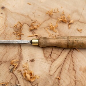 May include: A wooden handled chisel with a metal blade, lying on a wooden surface with wood shavings scattered around it.
