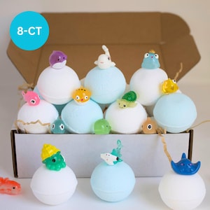 May include: An open cardboard box filled with bath bombs. The bath bombs are white and light blue, topped with small, colorful plastic sea creatures. A blue circle with the text "8-CT" is in the upper left corner.
