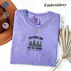 Könnte beinhalten: Ein fliederfarbenes Comfort Colors T-Shirt mit dem Text "COLUMBIA INN PINE TREE, VERMONT Always a White Christmas" in Schwarz und Grün. Eine goldene Halskette mit Anhänger liegt auf dem gefalteten Shirt.