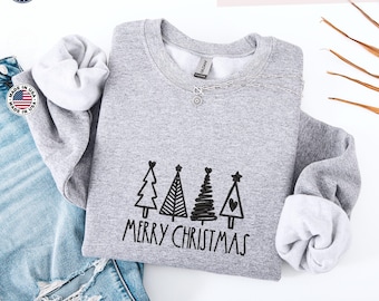 Può includere: Una felpa grigio chiaro con la parola "Ricamo" in alto. La felpa presenta un disegno ricamato nero di quattro alberi di Natale e le parole "BUON NATALE". Le maniche sono arrotolate e la felpa è abbinata a jeans blu.